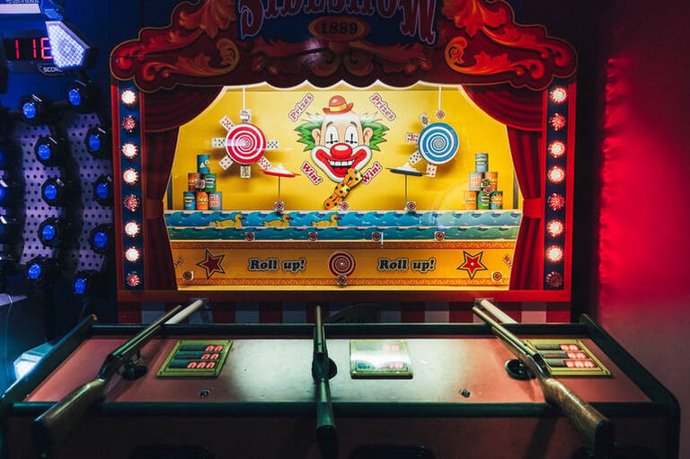 Vintage plinko-style game booth with colorful clown target and toy rifles under bright lights