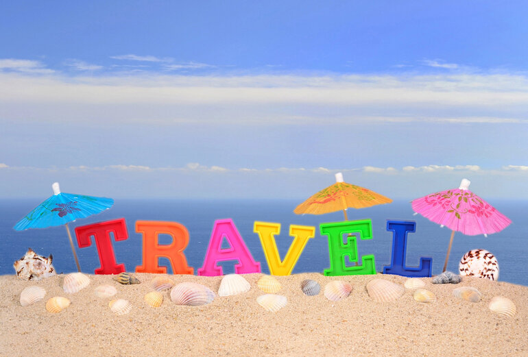 Colorful TRAVEL letters on a sandy beach with seashells, paper umbrellas, and a clear blue sky in the background