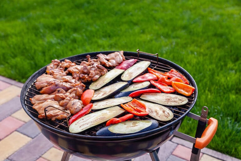 A charcoal grill in a backyard setting with chicken, eggplant, and bell peppers cooking on the grates