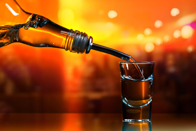 Close-up of alcohol being poured from a bottle into a shot glass against a warm, blurred background