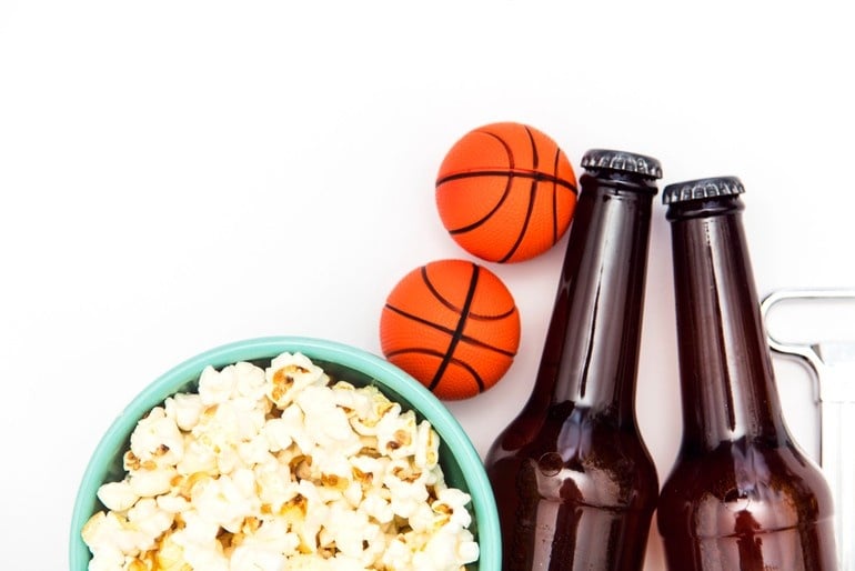 Flat lay of two craft beer bottles, a bowl of popcorn, and mini basketballs on a white background, representing game-day snacks and drinks