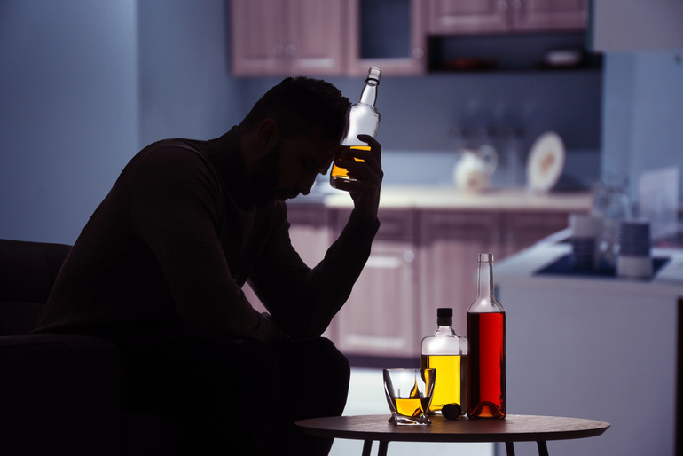 A distressed man sitting alone with bottles of alcohol on a table, suggesting emotional struggle and the potential influence of drinking on relationship decisions and infidelity