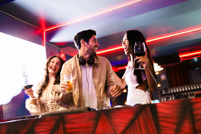 Three people smiling and holding drinks while standing near the DJ equipment in a bar with colorful neon lights
