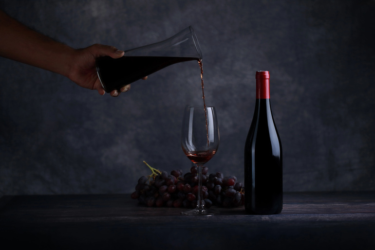 Hand pouring Pinot Noir into a liquor glass with bottle and grapes on a dark background