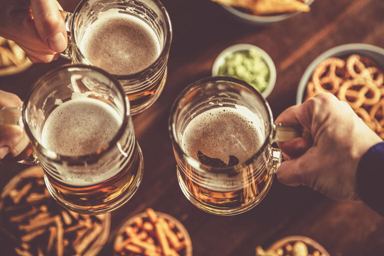 Three students clinking beer mugs over a table filled with snacks, capturing the social and cultural role of beer in college life