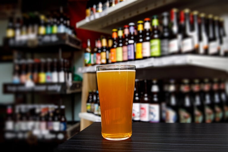 A pint of beer on a bar counter with shelves of bottled beers in the background