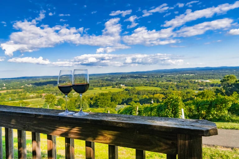 Two glasses of red wine on a wooden railing overlooking a sunny vineyard and rolling green hills under a bright blue sky in Arizona