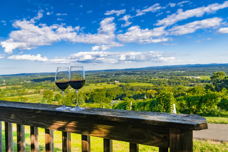 Two glasses of red wine on a wooden railing overlooking a sunny vineyard and rolling green hills under a bright blue sky in Arizona