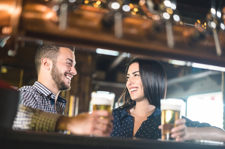 A man and woman smiling and holding glasses of beer while talking at a bar, creating a relaxed first-date atmosphere