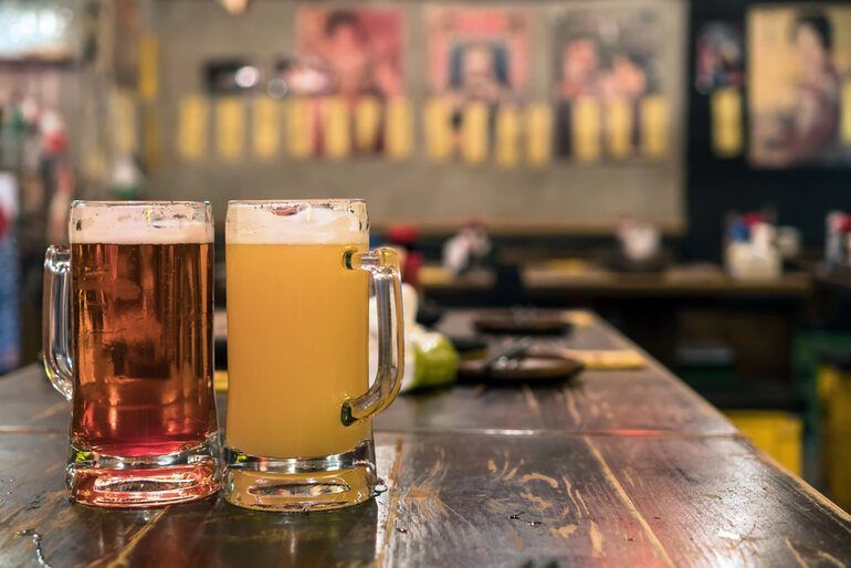 Two frosty beer mugs, one with amber ale, the other with hazy pale ale, on a rustic wooden table inside a cozy taproom