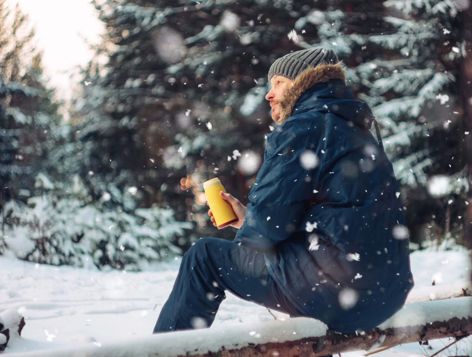 Man traveler forester hunter in winter forest sitting under snow on fallen tree with yellow thermos in his hands