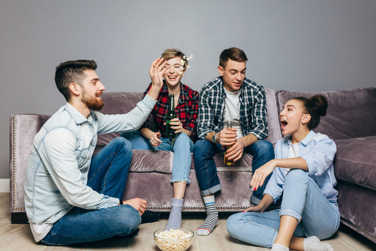 Friends having fun at home, drinking beer and eating popcorn during a movie night, laughing and enjoying time together on the couch.