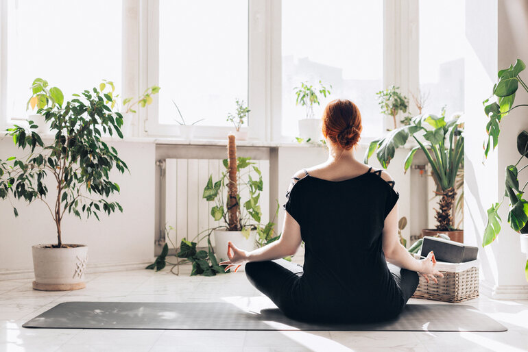 Woman practicing meditation on a yoga mat at home surrounded by indoor plants, focusing on mindfulness and present-moment grounding.