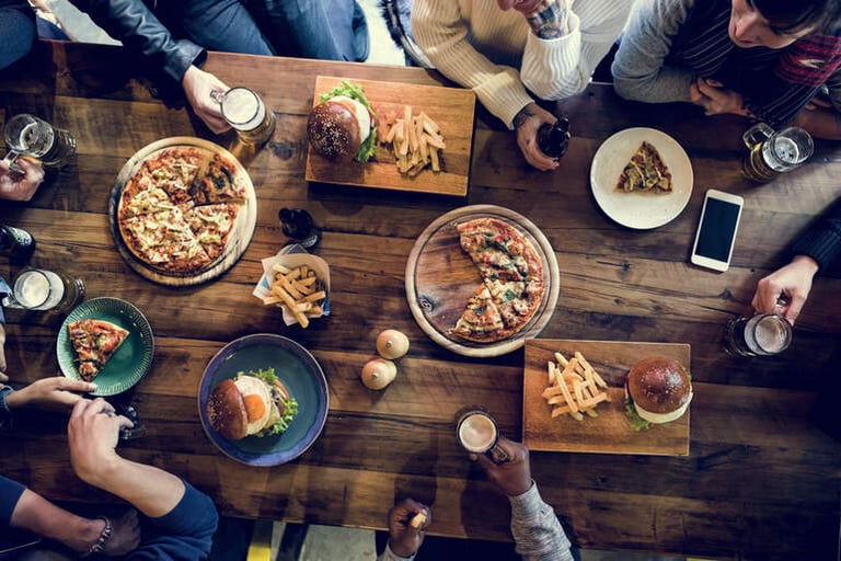 Group of people sharing pizzas, burgers, and drinks at a wooden table in a lively all-you-can-eat restaurant.
