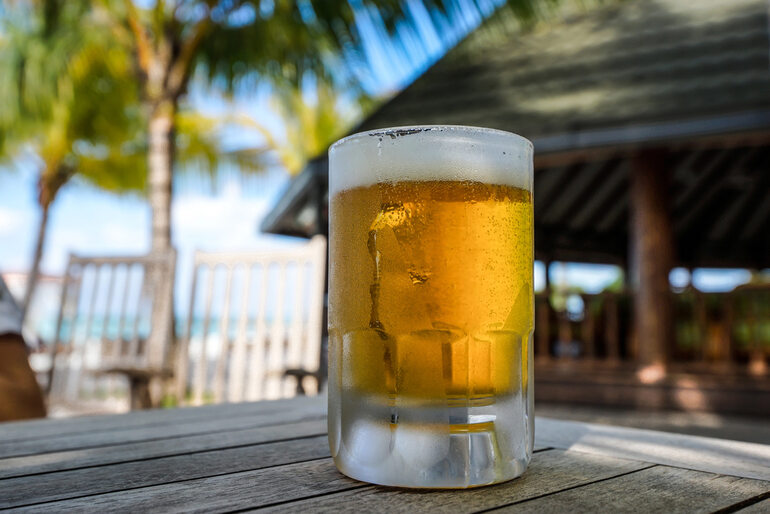 Cold Caribbean lager beer on a wooden table at the beach in the background with palm trees and the ocean.