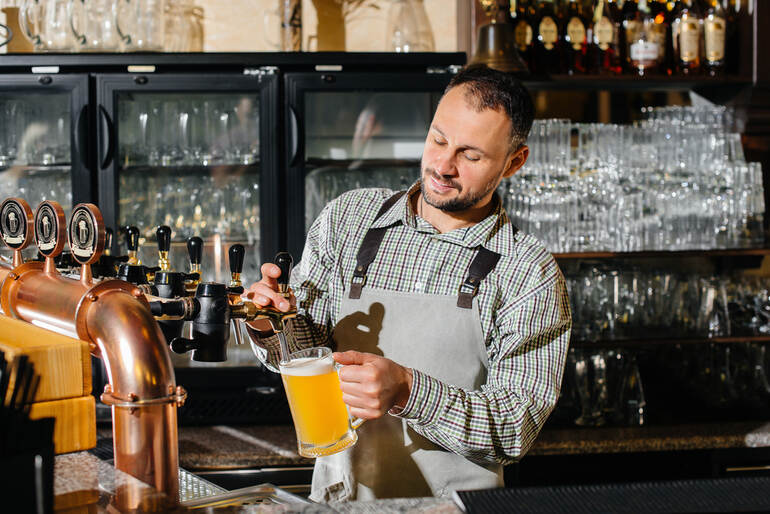 Bartender pouring a glass of craft beer from a tap inside a restaurant bar.