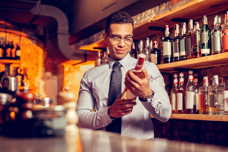 There is a male bartender who is examining a bottle of liquor at the counter of a full-stocked bar.