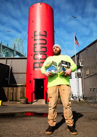 Rogue Ales brewer holding various six-packs in front of grain silo