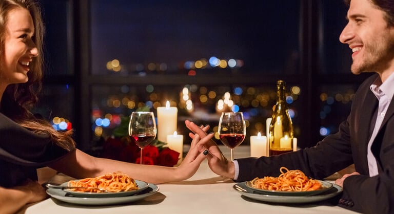 The couple is having a romantic Italian dinner with pasta, wine, and candlelight with a city view.
