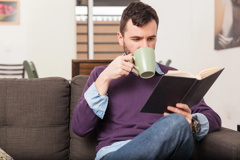 Man at home- relaxing as he reads a book- taking a hot non-alcoholic drink
