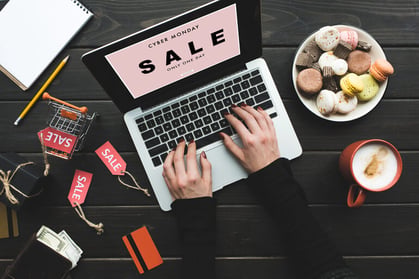 Person shopping online during a Cyber Monday sale, with laptop, credit card, shopping cart, and discount tags on desk