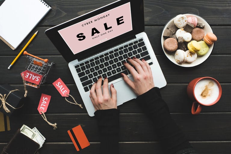 Person shopping online during a Cyber Monday sale, with laptop, credit card, shopping cart, and discount tags on desk