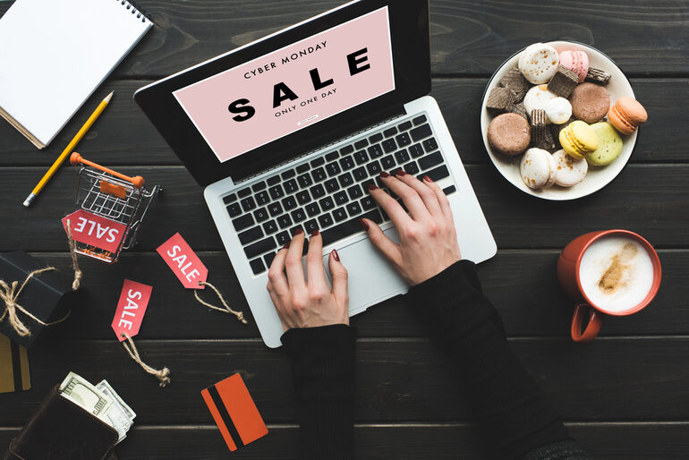 Person shopping online during a Cyber Monday sale, with laptop, credit card, shopping cart, and discount tags on desk