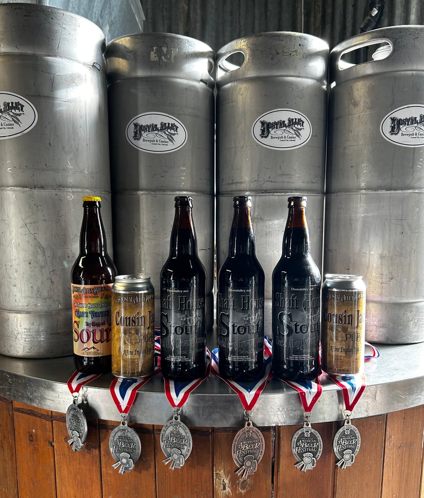 Dostal Alley beer bottles and cans displayed in front of stainless steel kegs, with award medals hanging below