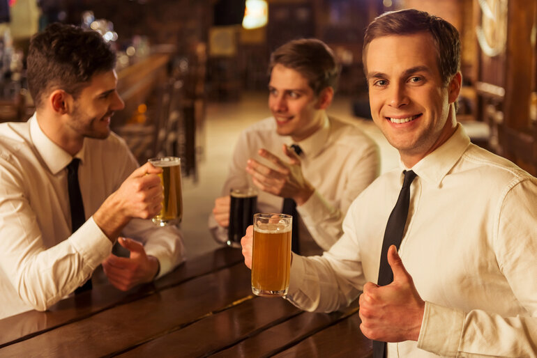 Three men in white shirts are sitting, smiling and holding mugs of beer. One man in the foreground is giving a thumbs-up