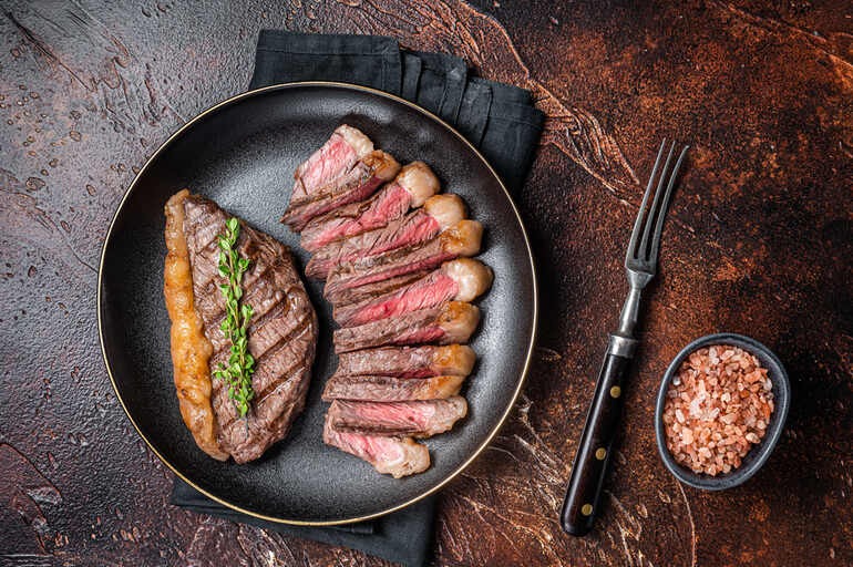 Sliced medium-rare picanha sirloin cap steak served on a black plate with a sprig of thyme, a fork, and coarse pink salt.