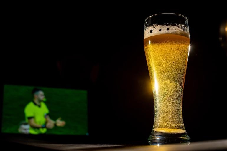 Glass of beer on a wooden shelf with a phone displaying a football match in the background.