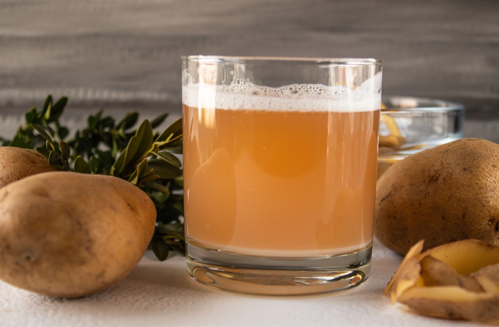 Glass of potato neutral spirit surrounded by raw potatoes and potato peels on a rustic table.