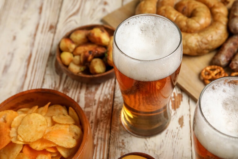 A cold glass of foamy beer surrounded by matchday snacks, including bowls of potato chips, roasted potatoes, pretzels, and grilled sausages on a wooden surface.