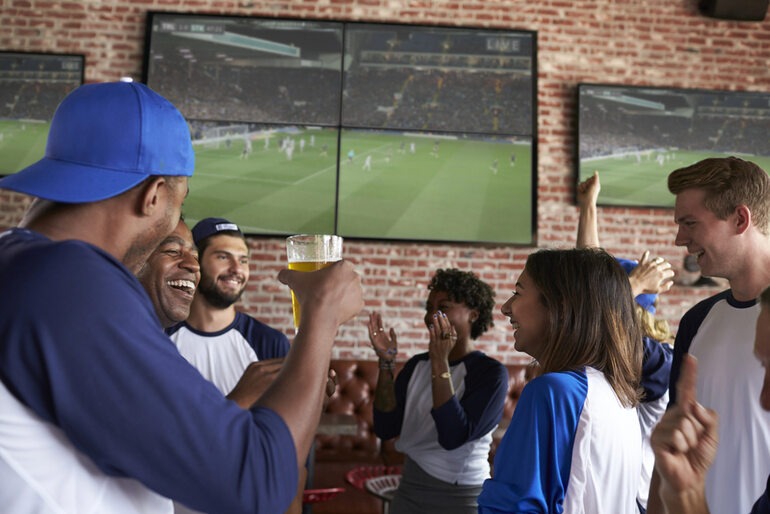 Friends cheering and enjoying beer while watching a match on screens in a lively taproom.