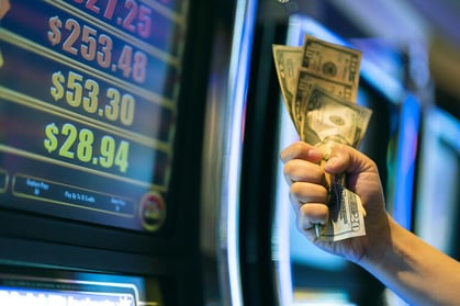 Person holding a stack of US dollar bills in front of a colorful slot machine display showing payout amounts for an article on slot paylines vs ways to win.