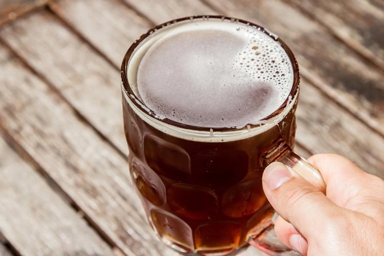 Hand holding a dimpled glass mug of dark craft beer on a wooden table, representing favorite beers in 'The Beer You Hunt And The Beer You Love Are Not The Same Beer'.
