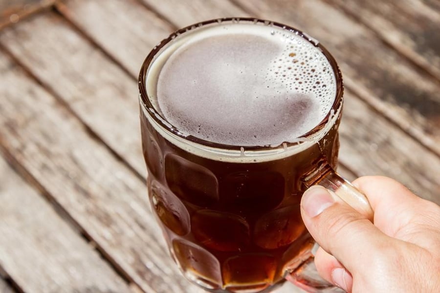 Hand holding a dimpled glass mug of dark craft beer on a wooden table, representing favorite beers in 'The Beer You Hunt And The Beer You Love Are Not The Same Beer'.