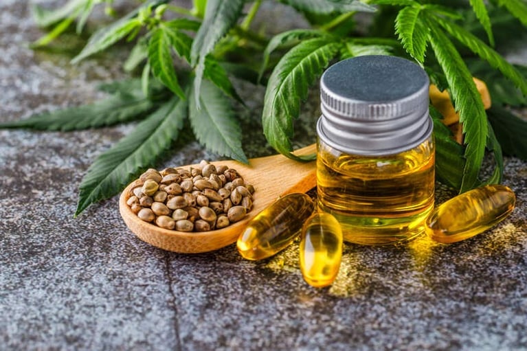 Small bottle of CBD oil with hemp seeds on a wooden spoon, amber capsules, and fresh leaves on a stone surface.