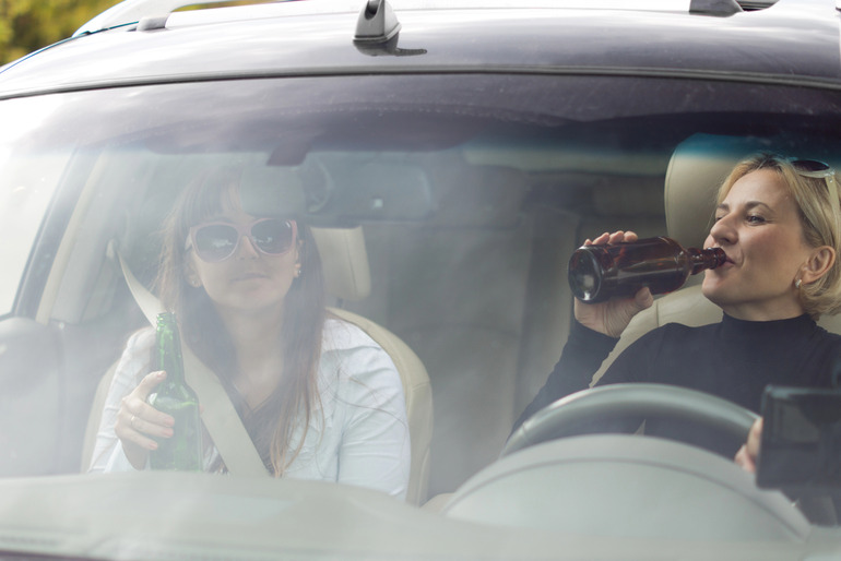 Two women drinking bottled beverages inside a vehicle during a brewery road trip.