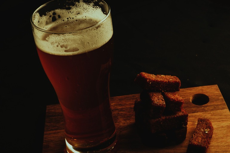 Dark beer with a foamy head served alongside toasted rye bread pieces on a wooden board.
