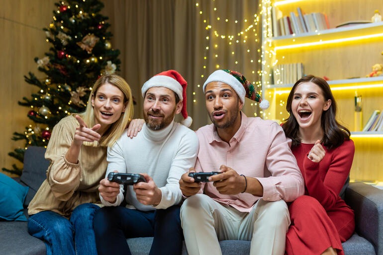 Friends wearing festive hats playing video games together on a couch in a Christmas-decorated living room during a holiday game night.