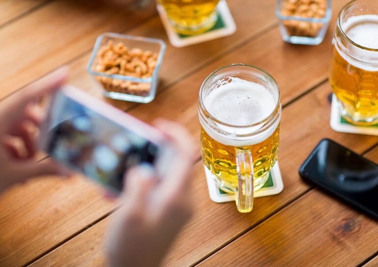 Person taking a photo of a glass of beer on a wooden table using a smartphone.