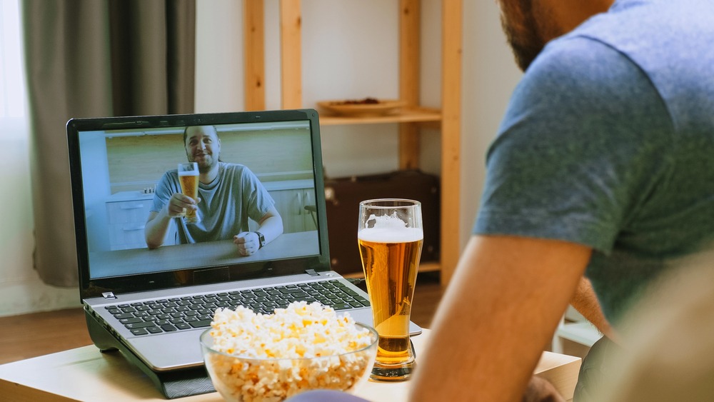 A man enjoying a virtual beer night with a friend over a laptop, with a glass of beer and popcorn on the table.